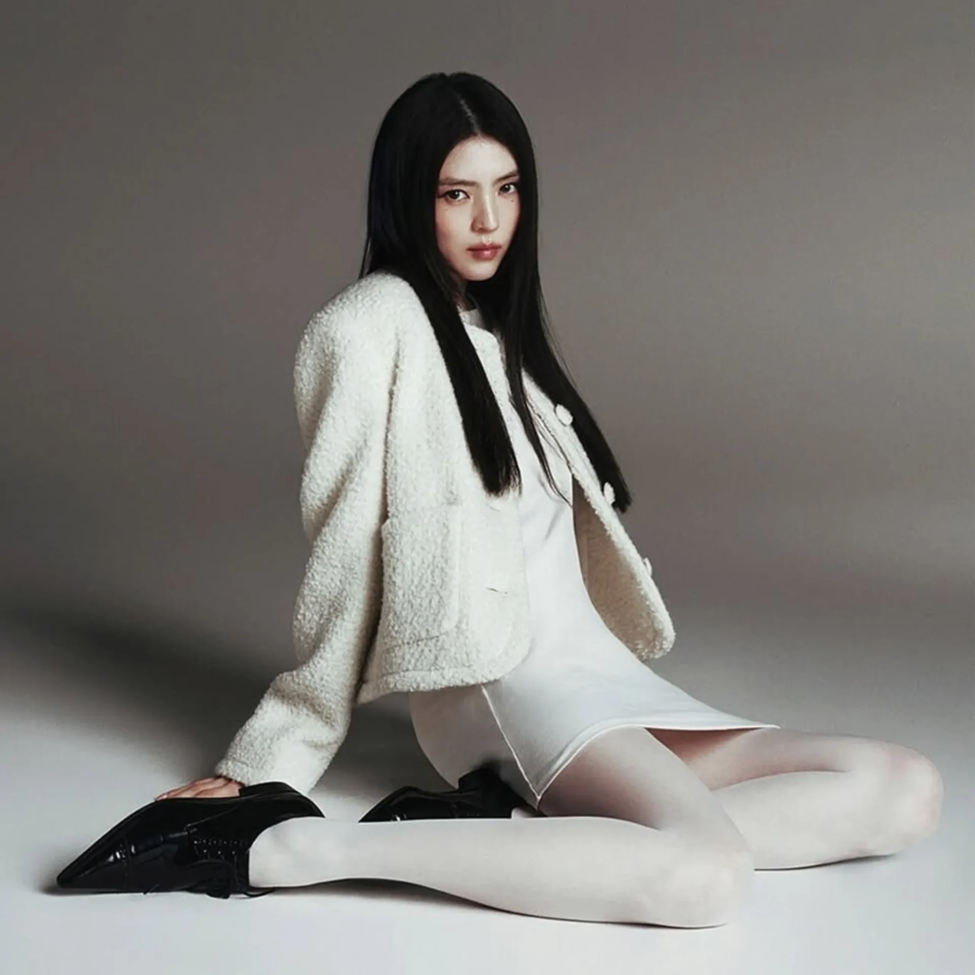 Woman with long dark hair in a white robe sits on a bed against a gray backdrop, looking at the camera.