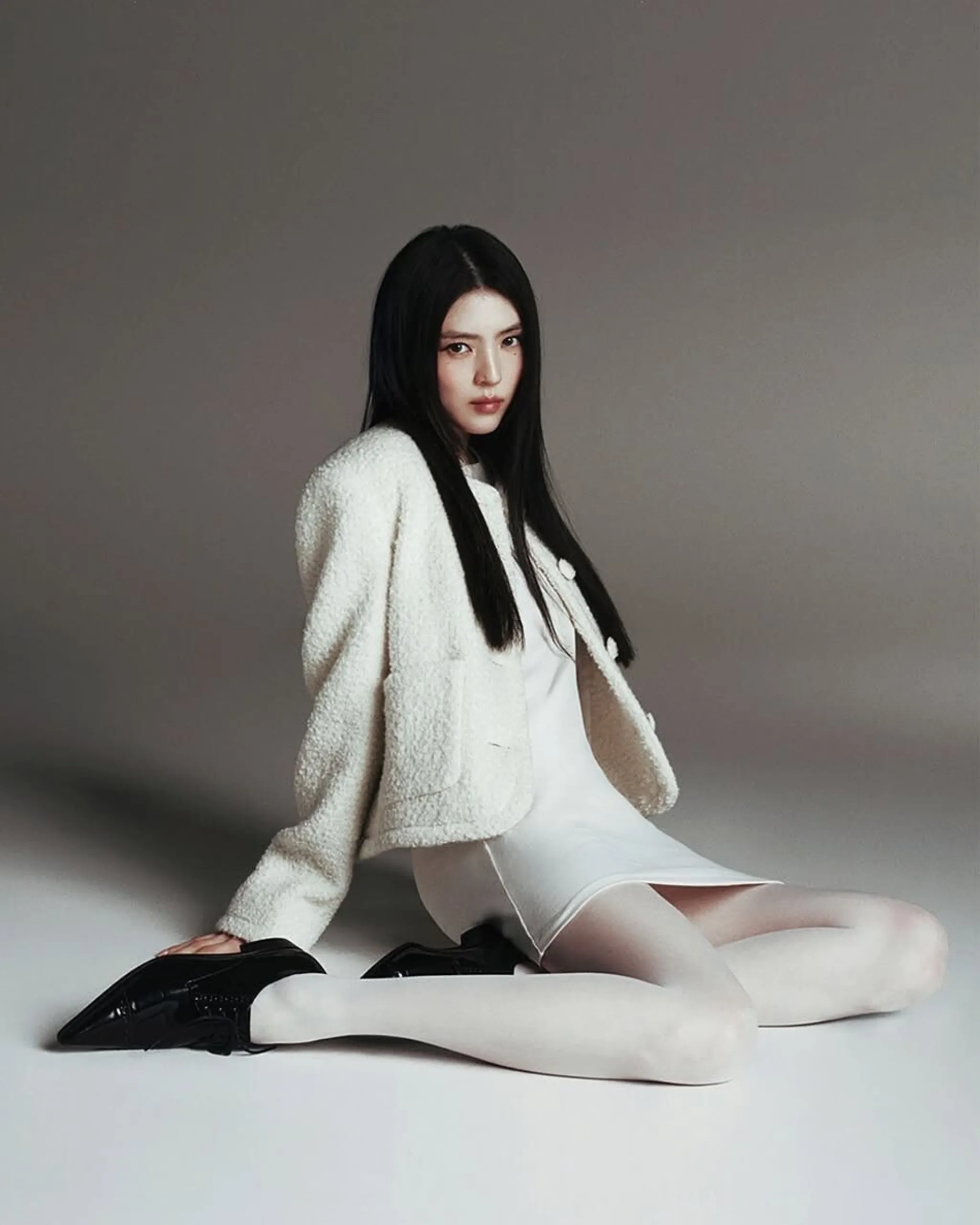 Model with long dark hair seated against a gray backdrop, wearing a white textured coat over a white dress and black shoes visible on the floor.