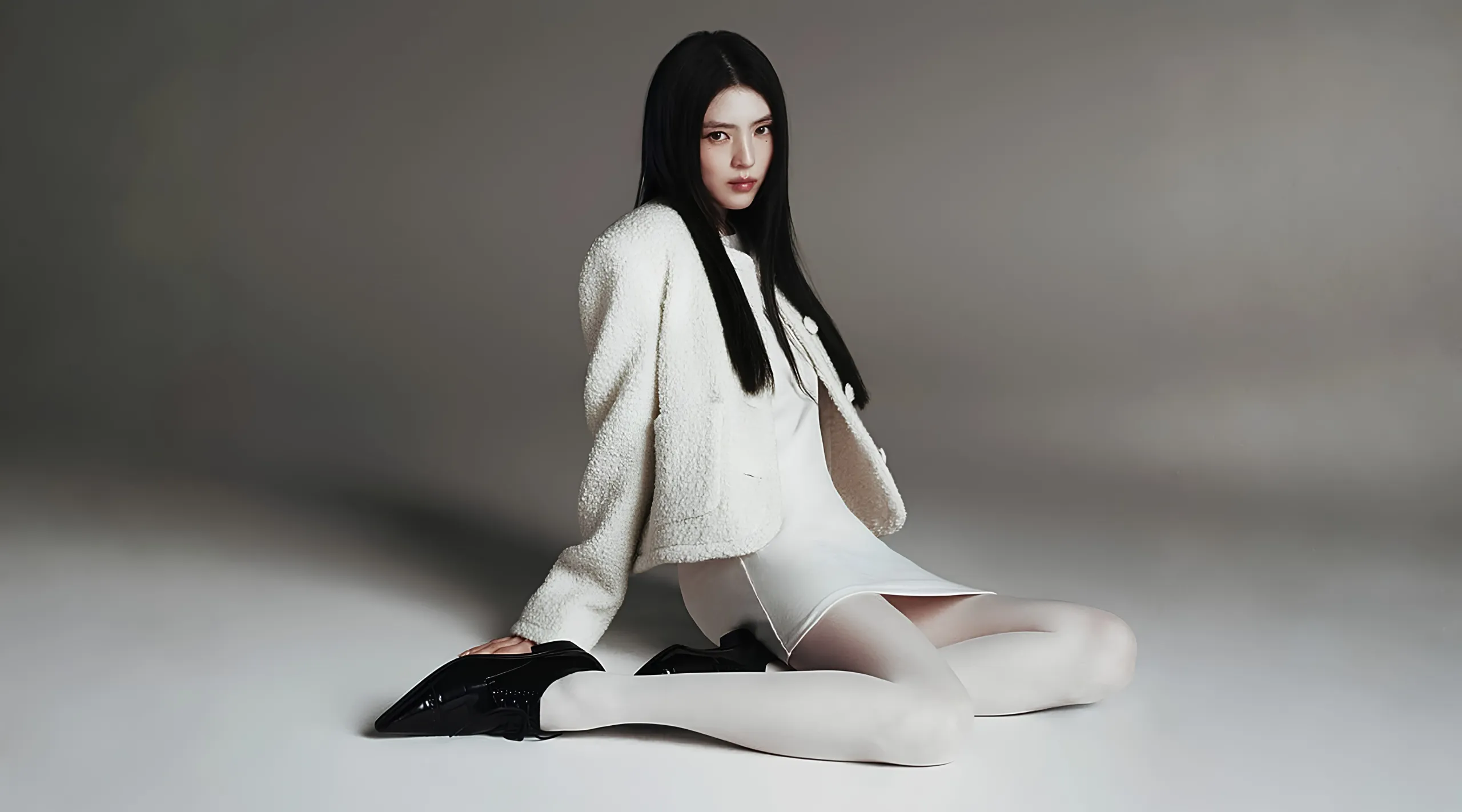 Model sits on the floor in a white textured coat and dress, with long black hair, against a neutral studio backdrop.