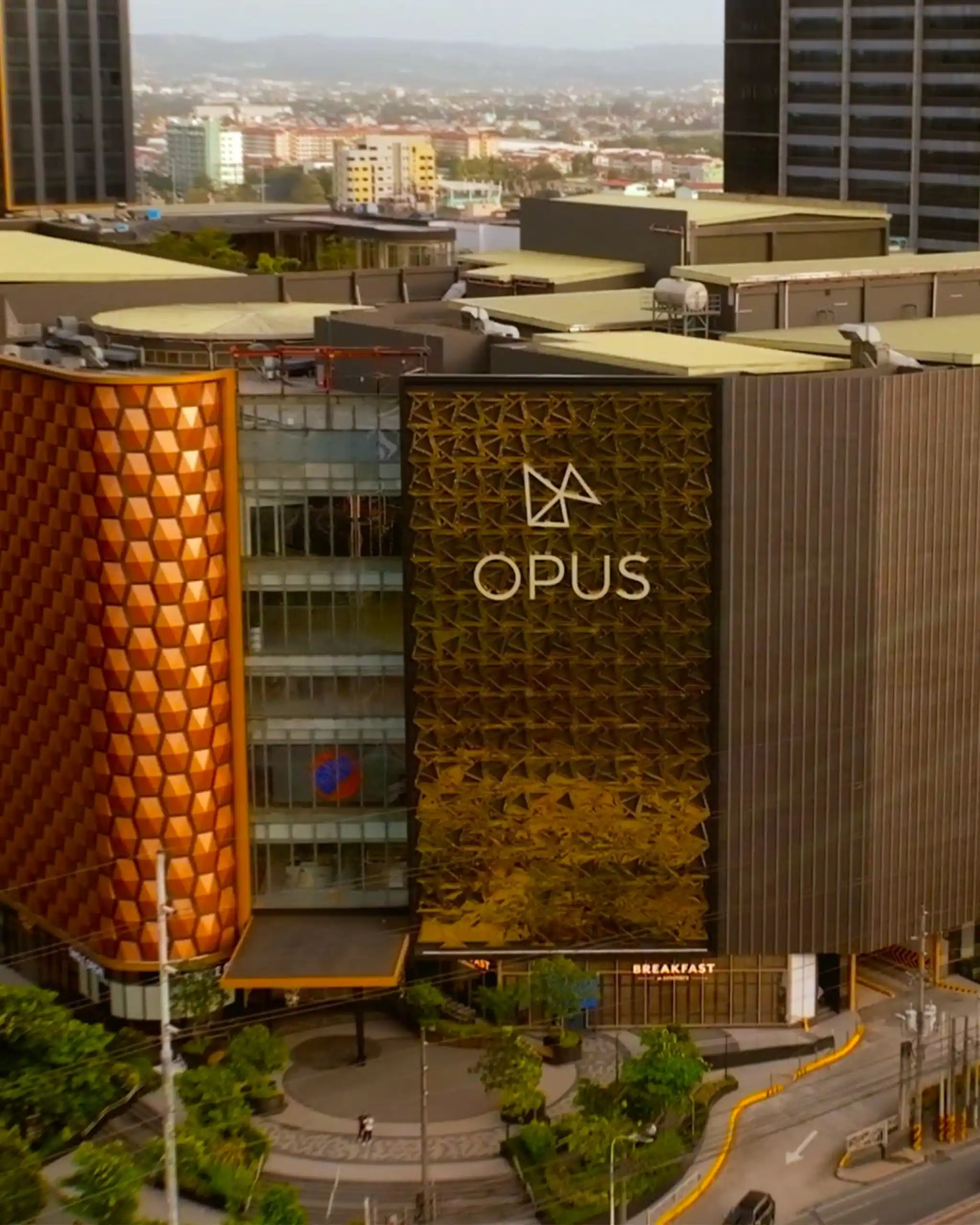 Tall building complex with orange geometric facade and a sign reading 'OPUS' on the front, overlooking a cityscape and curved road below.