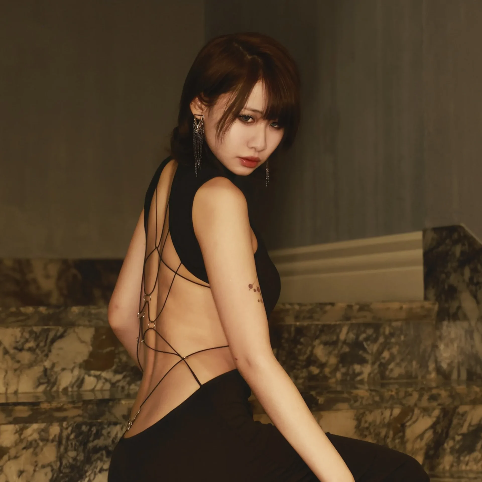 Woman in a black outfit turns toward the camera, posing against a dimly lit marble backdrop with dangling earrings. (Head and shoulders)