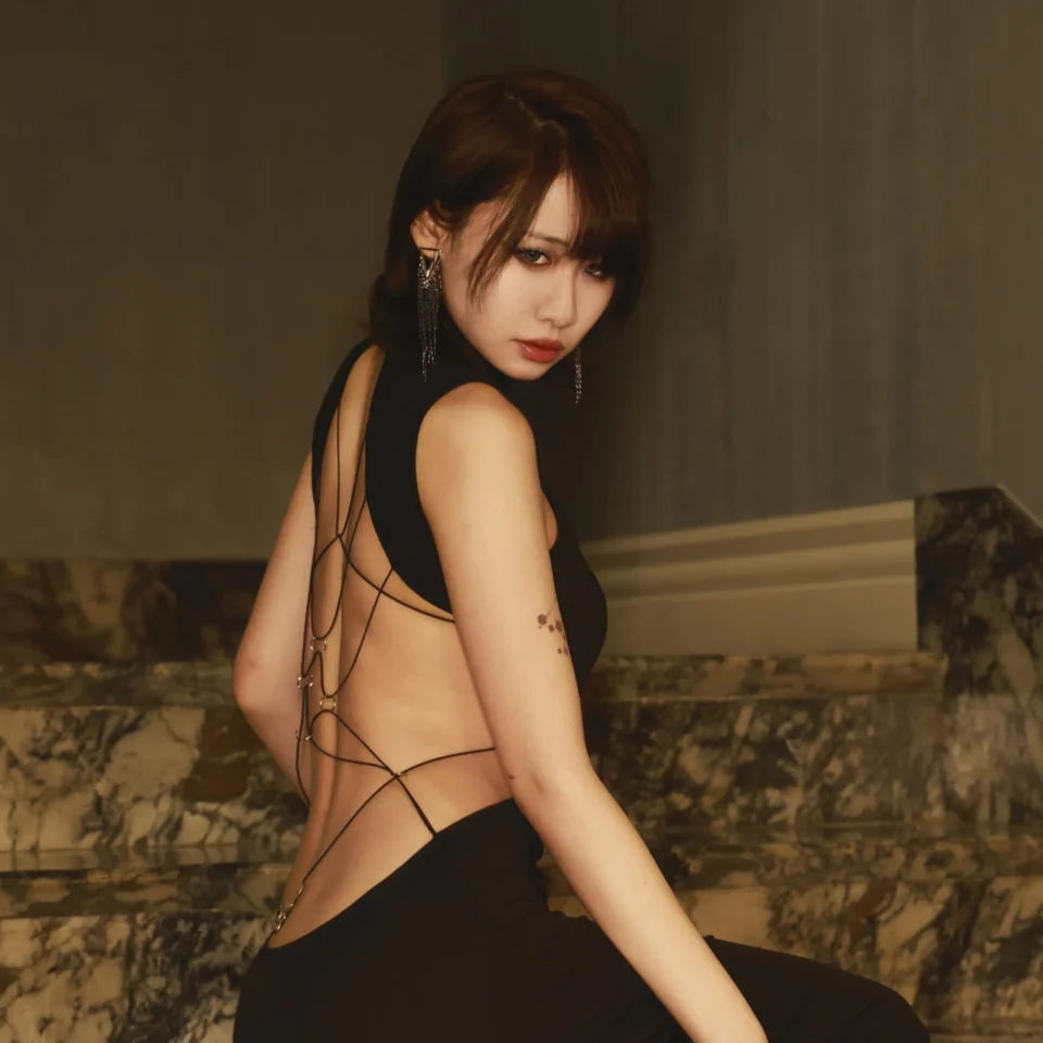 Woman in a black outfit turns toward the camera, posing against a dimly lit marble backdrop with dangling earrings. (Head and shoulders)