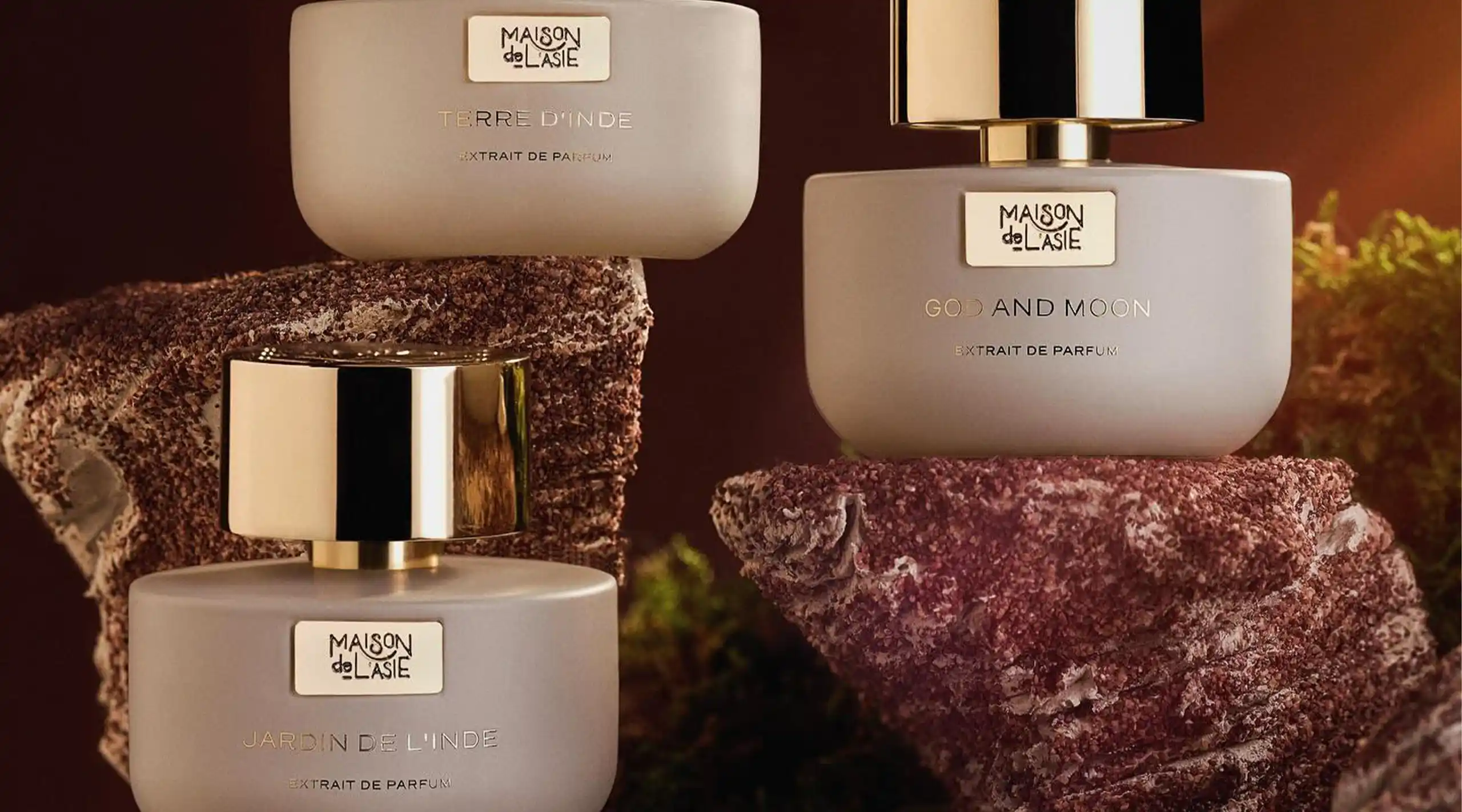 Three frosted perfume bottles with gold caps on pink stone pedestals; labels read Jardin de l’Inde and Terre d’Inde (Extrait de Parfum).