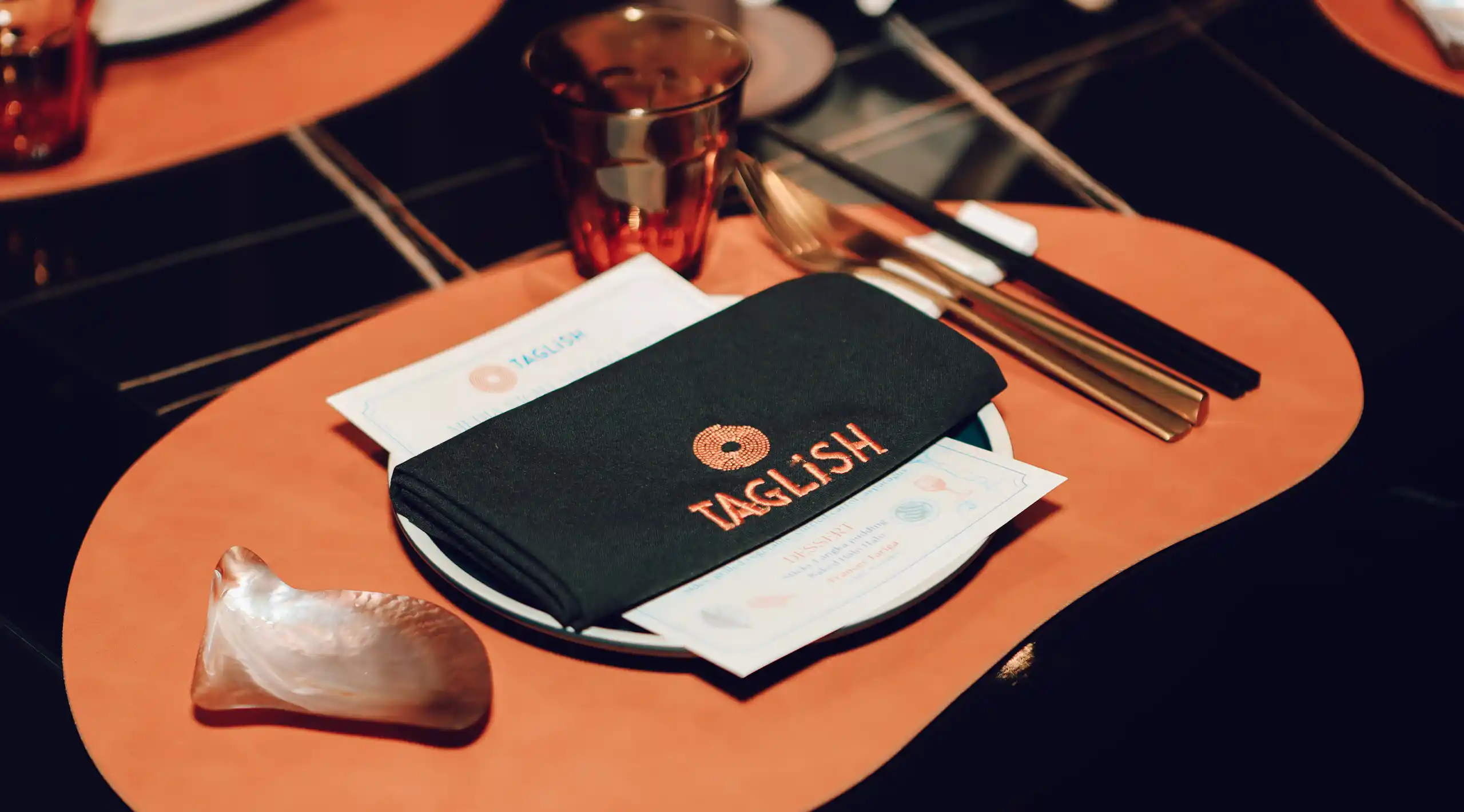 Table setting with orange placemat, a black napkin embroidered with TAGLISH, a glass, cutlery, and menus underneath the napkin.