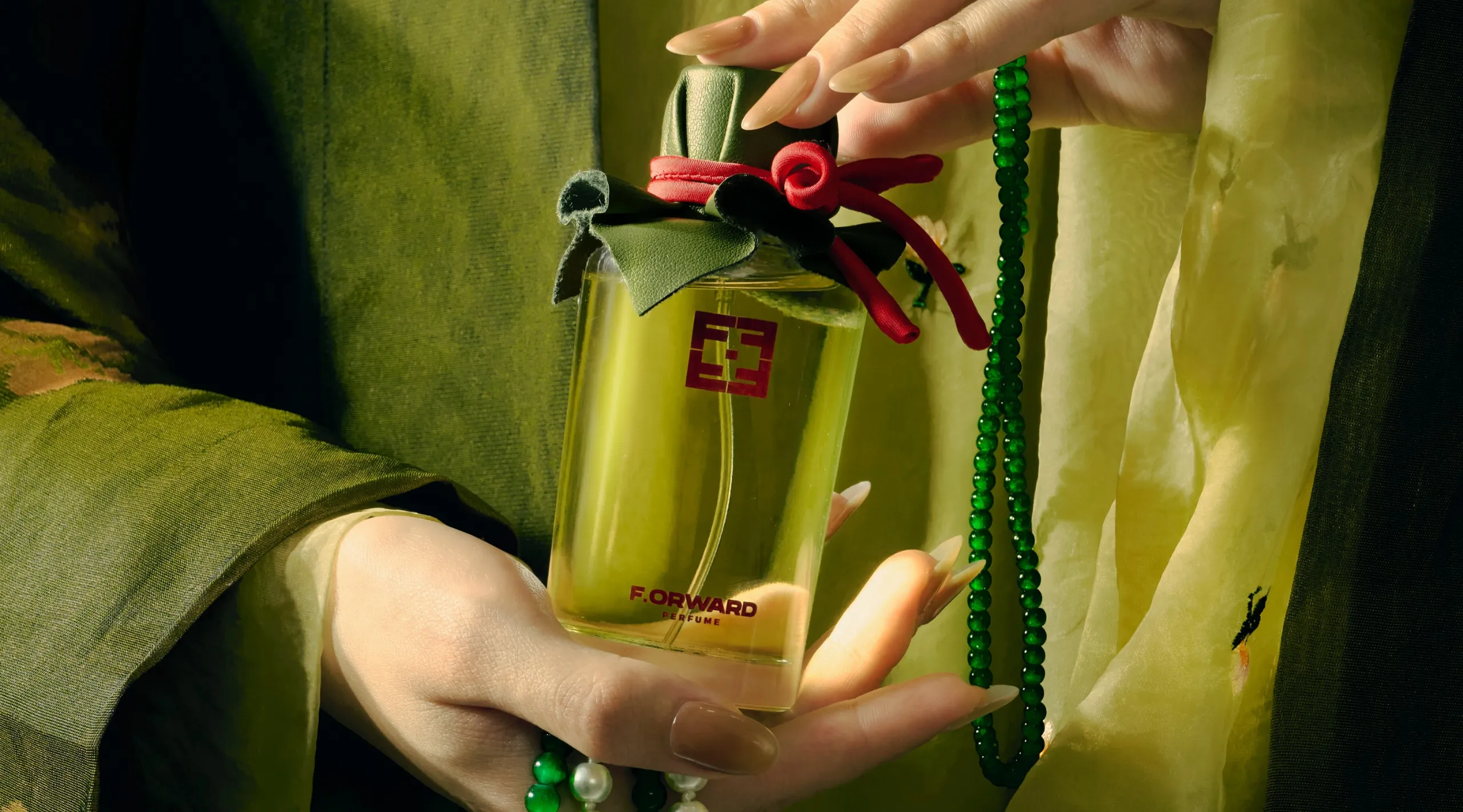 Hand holding a bottle of green perfume with a dark green velvet cap, red ribbon, and green bead necklace draped around the bottle.