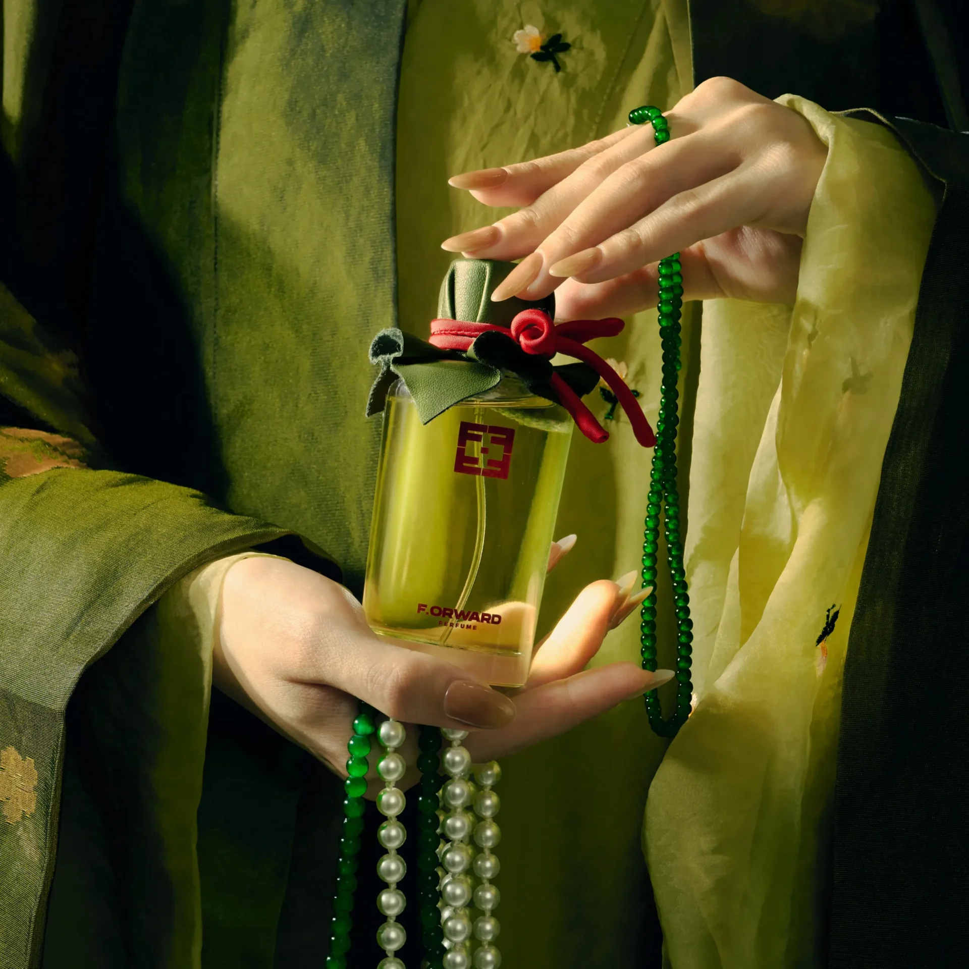 Close-up of a pale hand with long manicured nails holding a transparent perfume bottle tied with green and red ribbons, with green and pearl bead necklaces draped over the wrist; bottle label reads “F. Forward Perfume.”