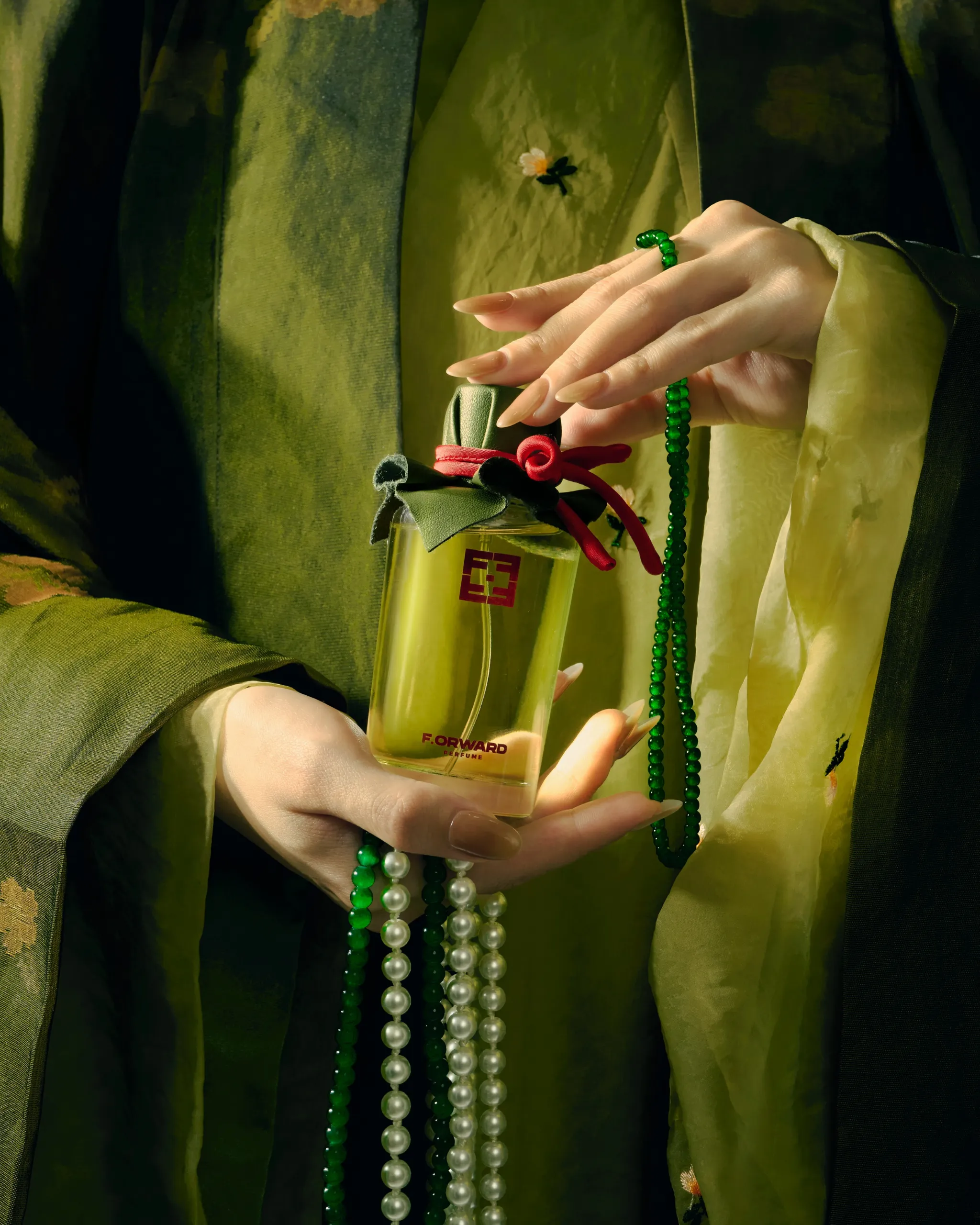 Hand with nude nails holding a glass perfume bottle wrapped in a green ribbon and red bow, with green bead jewelry against a green fabric backdrop.