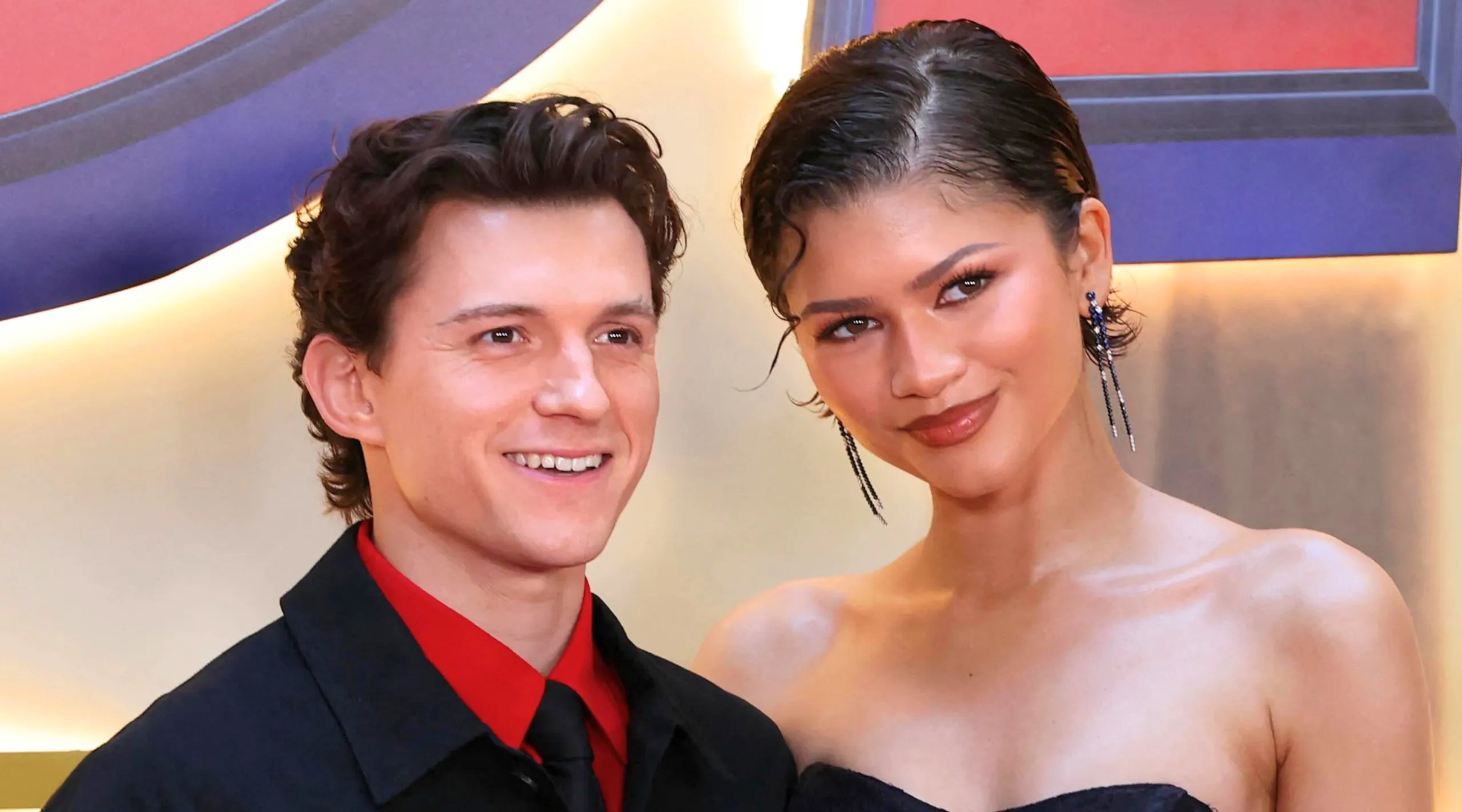 Smiling man in a black suit with a red shirt posing next to a woman in a black strapless dress on a red carpet backdrop.