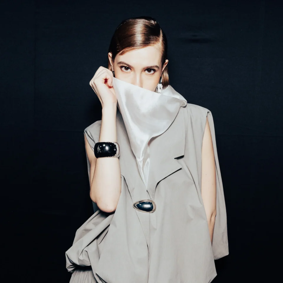 Person with slicked-back hair wearing a large gray collar, partially covering their face against a dark background.