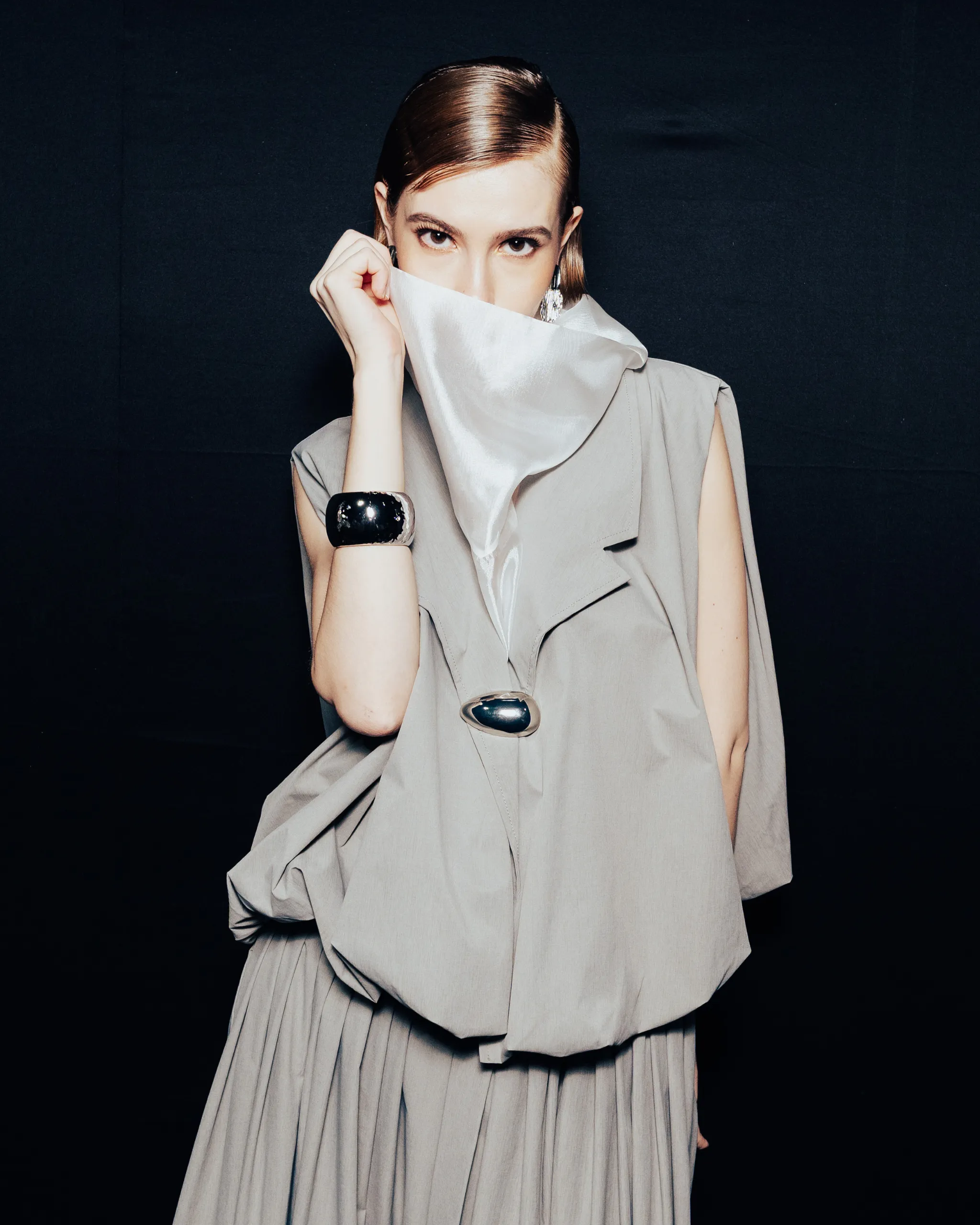 Woman in a gray, structured dress covers her mouth with a white scarf, posed against a dark backdrop.