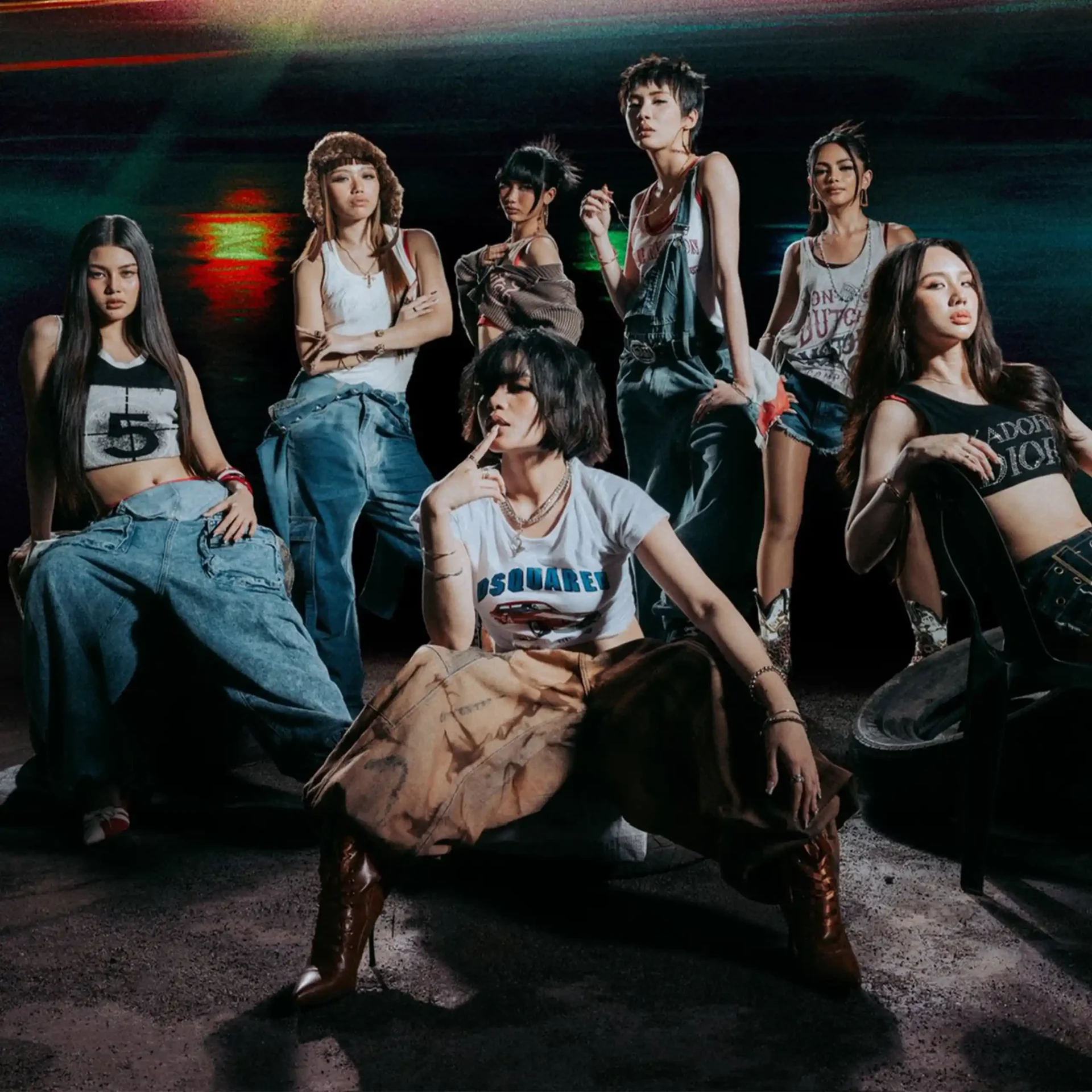Group of stylish young women posing together in casual streetwear (crop tops and denim) in a dark, urban setting with colored lighting in the background.