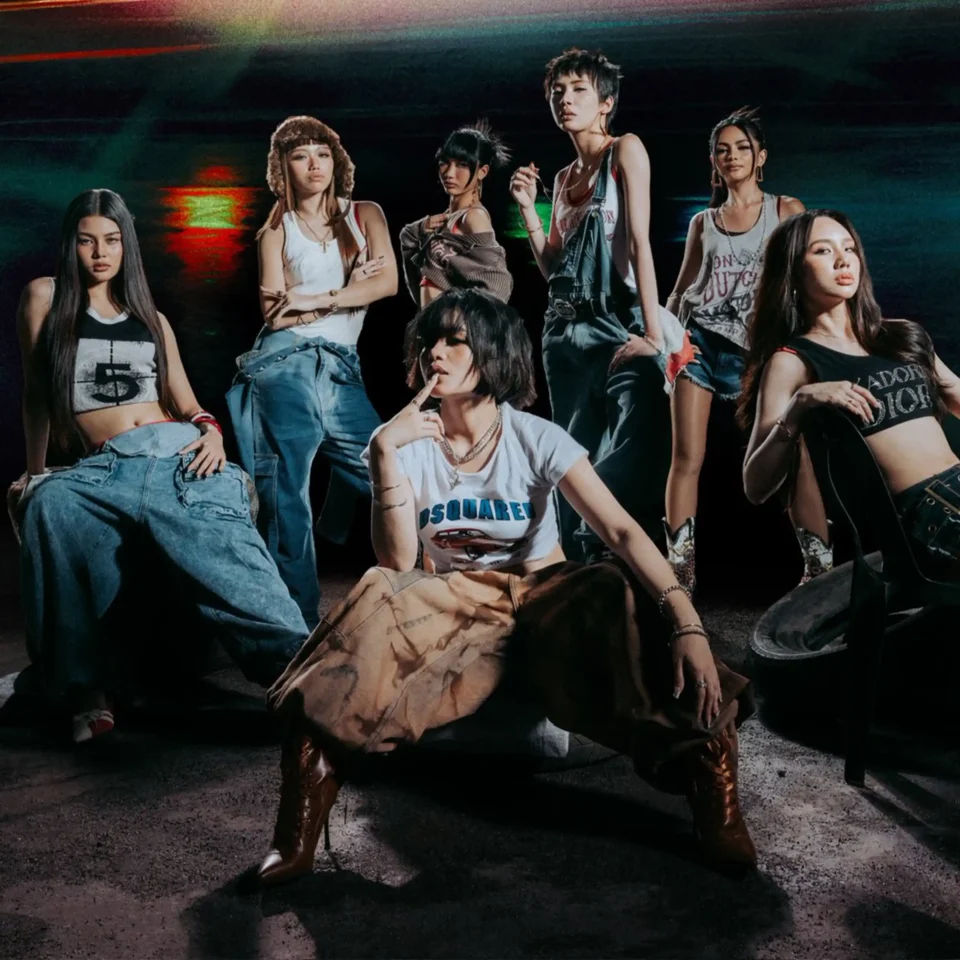 Group of stylish young women posing together in casual streetwear (crop tops and denim) in a dark, urban setting with colored lighting in the background.